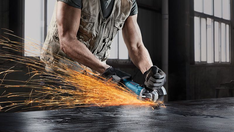 A person wearing safety equipment grinds metal using an angle grinder, sparks flying.