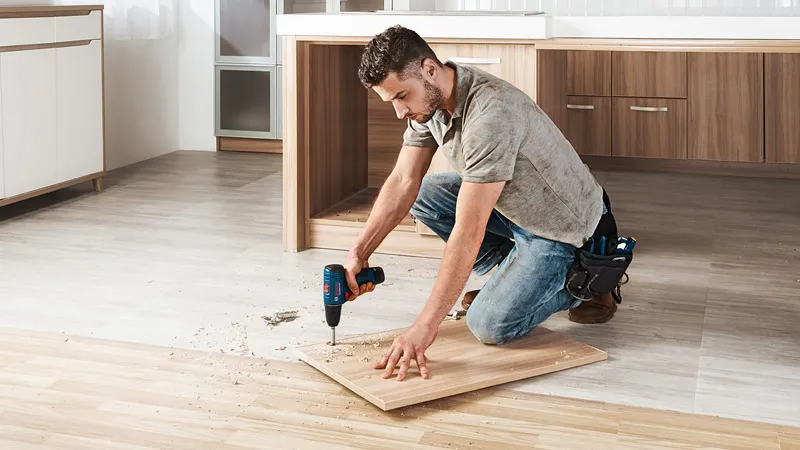 A person drills into a wooden board on the floor in a kitchen under renovation.