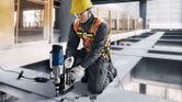 A person wearing safety equipment drills into steel beams at a construction site.