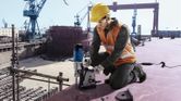 A person wearing safety equipment operates a magnetic drill on a shipyard platform.