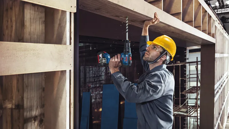 A person wearing safety equipment drills into a wooden beam at a construction site.