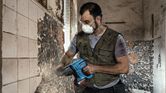 A person wearing safety equipment removes tiles from a wall using a power tool.