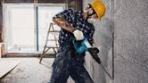 A person wearing safety equipment uses a demolition hammer to break a concrete wall.