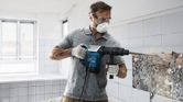 A person wearing safety equipment uses a demolition hammer to break wall tiles.