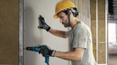 Person wearing safety equipment uses a drywall screwdriver to fasten a panel to a wall.