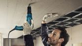 A person wearing safety equipment secures drywall to a ceiling using a screwdriver.