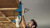 A person wearing safety equipment drives screws into a ceiling with a drywall screwdriver.