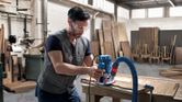 Person wearing safety equipment uses a router to shape wood in a workshop.