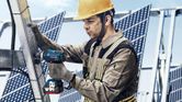 A person wearing safety equipment uses a cordless impact wrench on a metal solar panel frame.