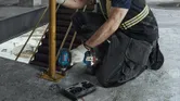 A person wearing safety equipment tightens a bolt on a metal post using a cordless impact wrench.