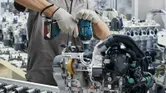 A person wearing safety equipment uses a cordless impact wrench on an engine assembly.