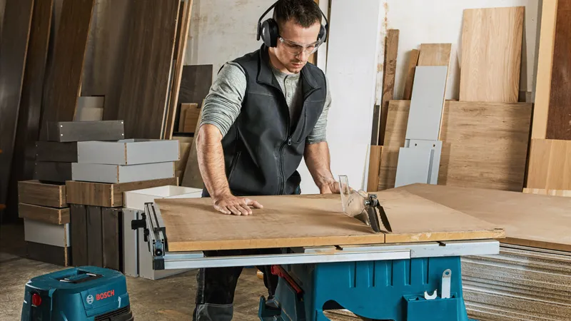 A person wearing safety equipment cuts wood using a table saw in a workshop.