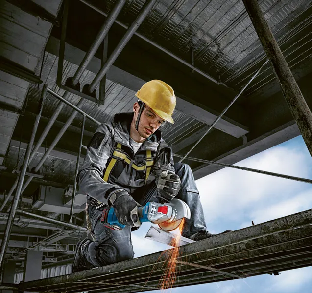 A person wearing safety equipment uses a cordless angle grinder to cut metal.
