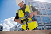 A person wearing safety equipment drills a large hole in wood with a cordless tool.