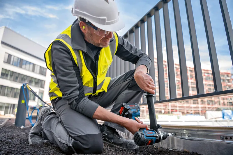 A person wearing safety equipment uses a cordless combi drill on a metal fence frame.