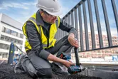 A person wearing safety equipment uses a cordless combi drill on a metal fence frame.