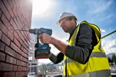 A person wearing safety equipment drills into a brick wall with a cordless combi tool.