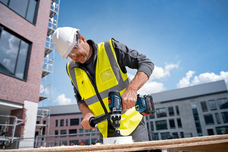 A person wearing safety equipment drills a hole into wood at a construction site.