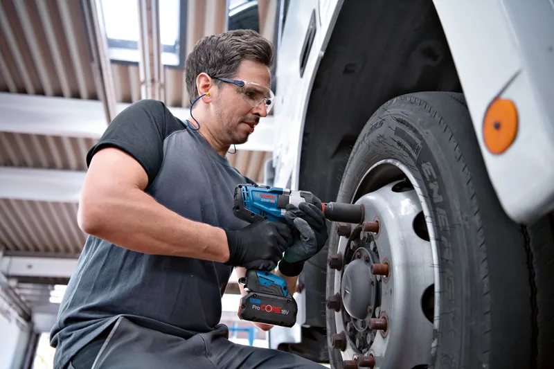 A person wearing safety equipment uses a cordless impact wrench to tighten truck wheel bolts.