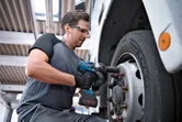 A person wearing safety equipment uses a cordless impact wrench to tighten truck wheel bolts.