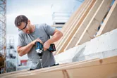 Person wearing safety equipment uses a cordless impact wrench to drive a screw into wood.