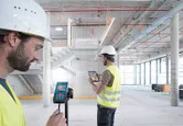 Two persons wearing safety equipment use a laser measure and a tablet inside a building under construction.