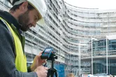 A person wearing safety equipment operates a laser measure at a construction site.