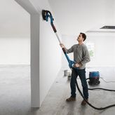 A person sands a white wall with a drywall sander connected to a dust extractor.