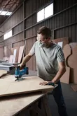 A person wearing safety equipment cuts plywood with a cordless jigsaw in a workshop.