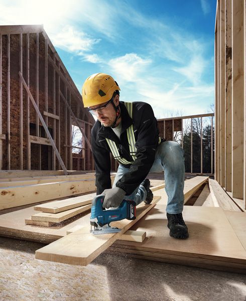 GST 185-LI A person wearing safety equipment cuts a wooden plank with a cordless jigsaw at a construction site.