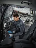 A person wearing safety equipment uses a power tool to assemble a car door on a factory line.