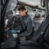 A person wearing safety equipment uses a power tool to assemble a car door on a factory line.