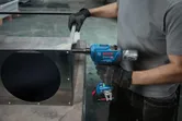 A person wearing safety equipment uses a cordless impact wrench on a metal duct.