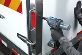 A person wearing safety equipment uses an impact wrench to secure a bolt on a metal door.