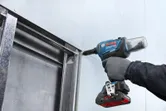 A person wearing safety equipment fastens drywall to a metal frame with a power tool.