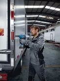 A person wearing safety equipment uses a cordless rivet gun on a truck door.