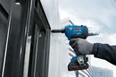 A person wearing safety equipment fastens a metal panel with a cordless rivet gun.