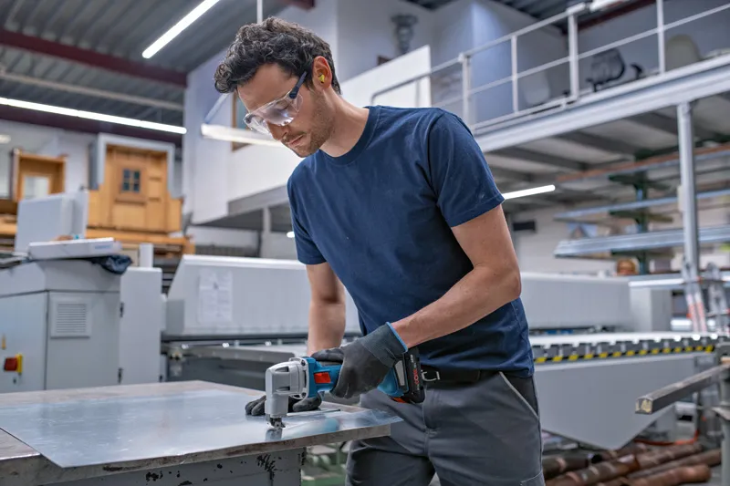 A person wearing safety equipment cuts sheet metal with a cordless metal shear.