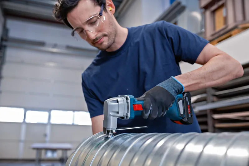 A person wearing safety equipment cuts a metal duct with a cordless metal shear.