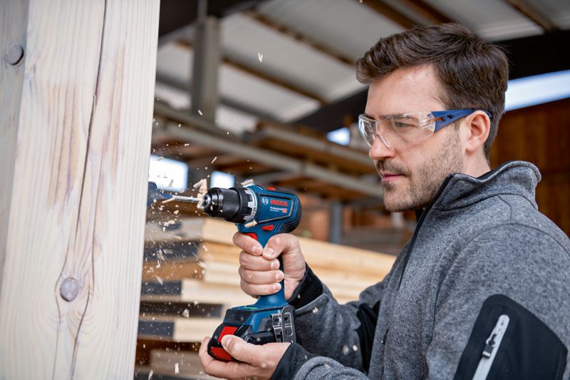 Person wearing safety equipment drills into a wooden beam in a workshop.
