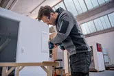 A person wearing safety equipment drills a hole in wood using a cordless power drill.