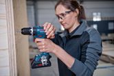 A person wearing safety equipment uses a cordless drill on a wooden panel.