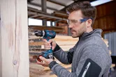A person wearing safety equipment drills into a wooden beam in a workshop.