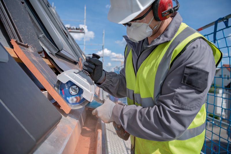 A person wearing safety equipment uses a cordless angle grinder to cut roof tiles.