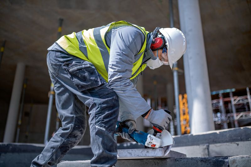 A person wearing safety equipment uses a cordless angle grinder to cut a concrete slab.