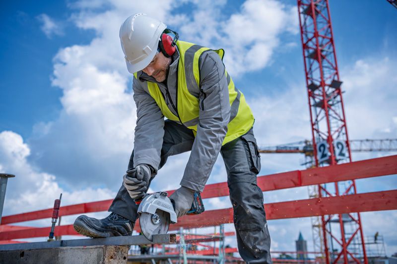 A person wearing safety equipment cuts metal on a construction site with a cordless angle grinder.