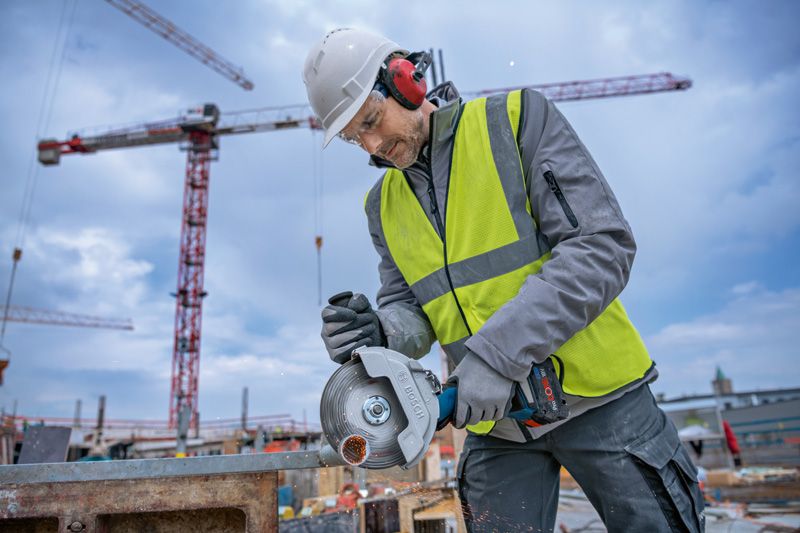 A person wearing safety equipment uses a cordless angle grinder to cut metal at a construction site.