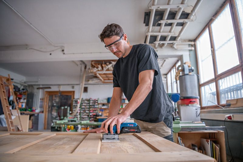 A person wearing safety equipment sands a wooden door with a cordless orbital sander.