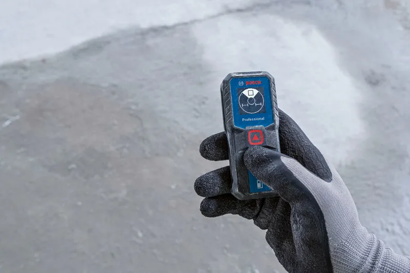 A person wearing safety equipment operates a laser measure on a concrete surface.