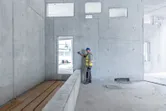 A person wearing safety equipment uses a laser measure against a concrete wall.